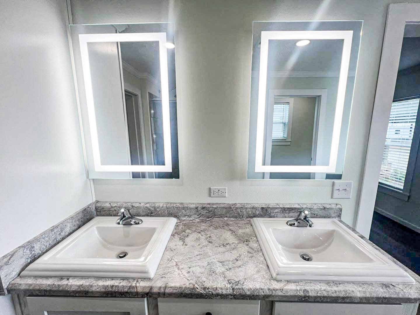 Modern bathroom with two white sinks set in a granite countertop, under sleek, illuminated mirrors. The ambiance is clean and contemporary.