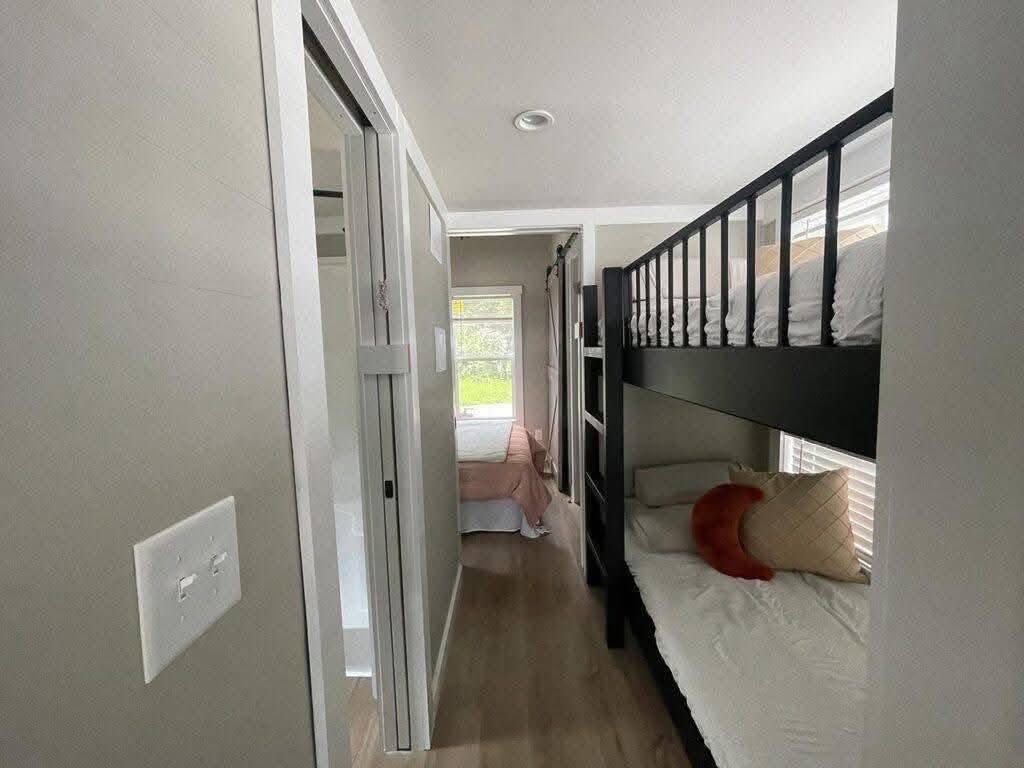 Narrow room with dark wood bunk beds on the right, white bedding and pillows. A hallway leads to a room with a bed and window, creating a cozy atmosphere.
