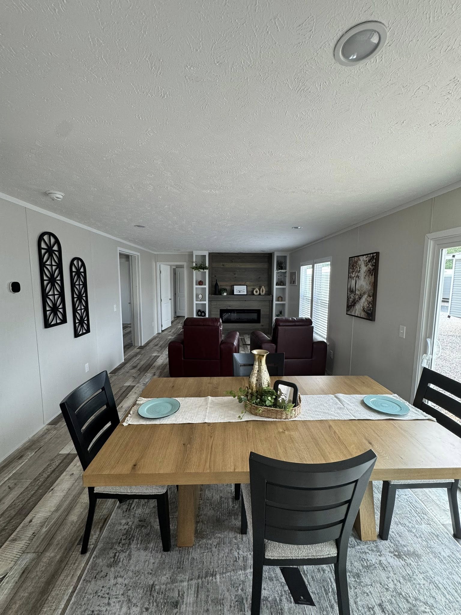 Modern living and dining area with a wooden table set for two, featuring turquoise plates and a decorative centerpiece. In the background, red armchairs face a fireplace with built-in shelves. The room is bright and airy, with light gray walls and a cozy ambiance.