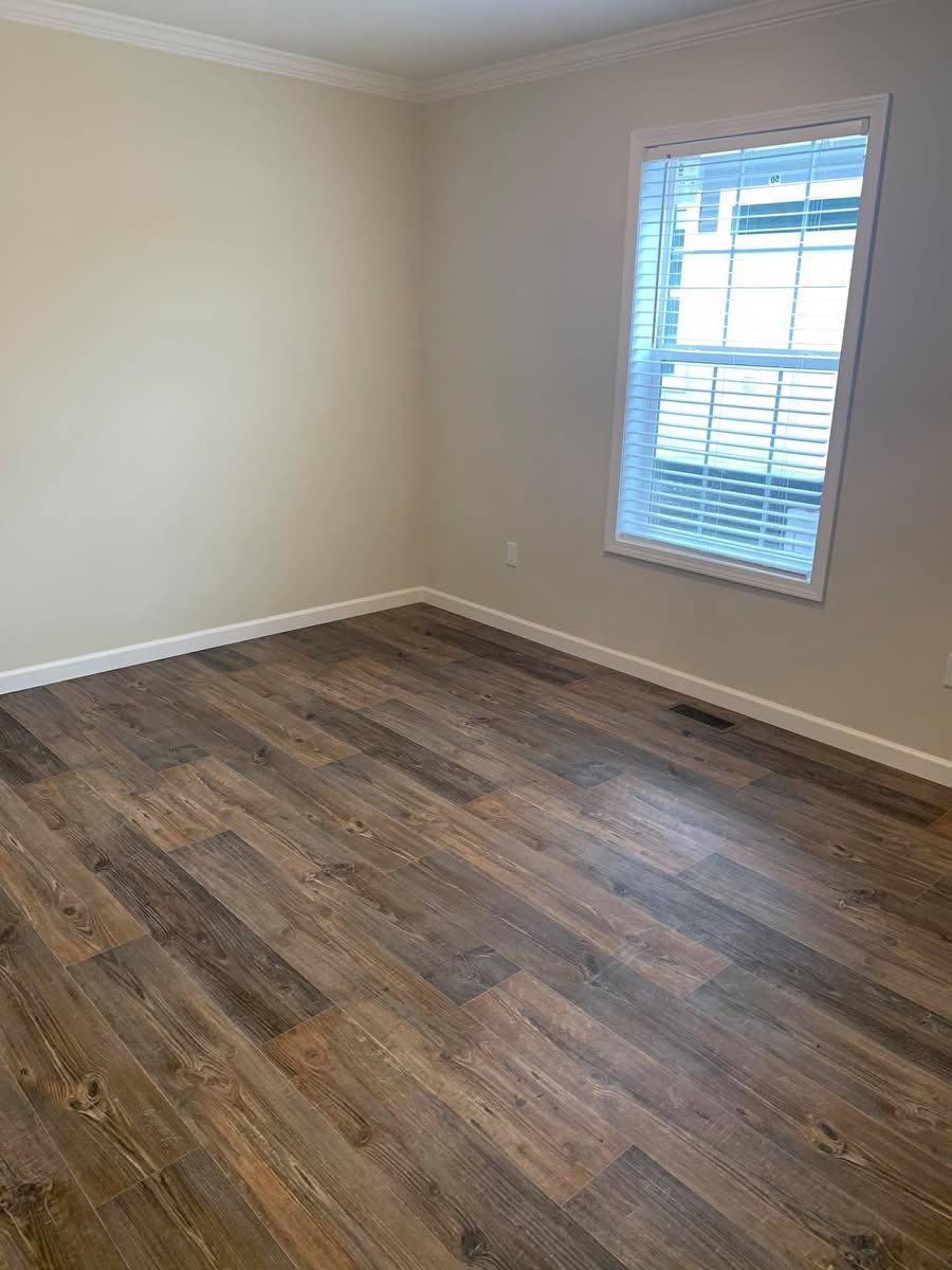 An empty room with light beige walls and dark wood laminate flooring. A large window with white blinds lets in natural light, creating an airy feel.