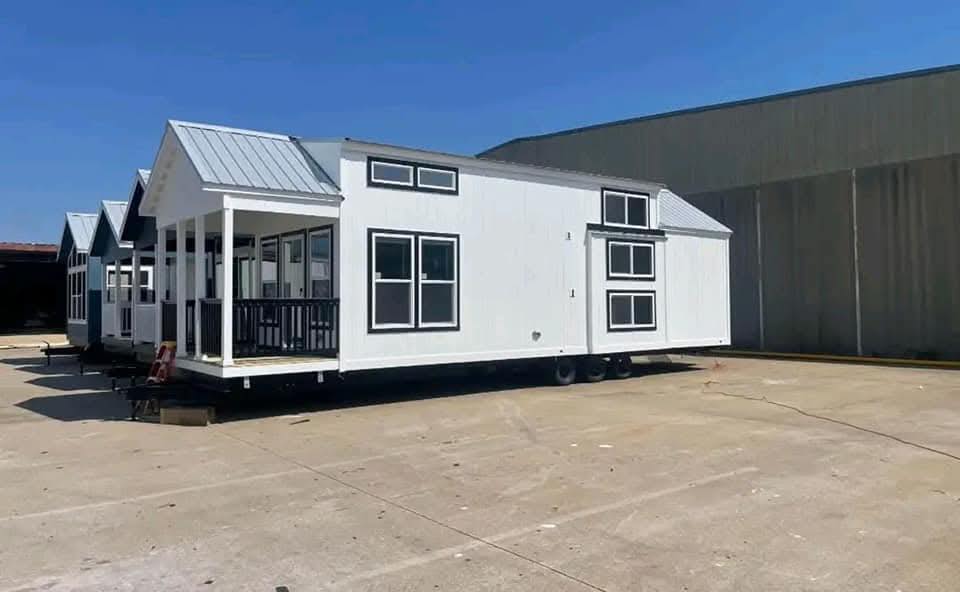A modern tiny house with white siding and black trim sits on a trailer. It features large windows, a small porch, and a pitched roof, exuding a cozy feel.