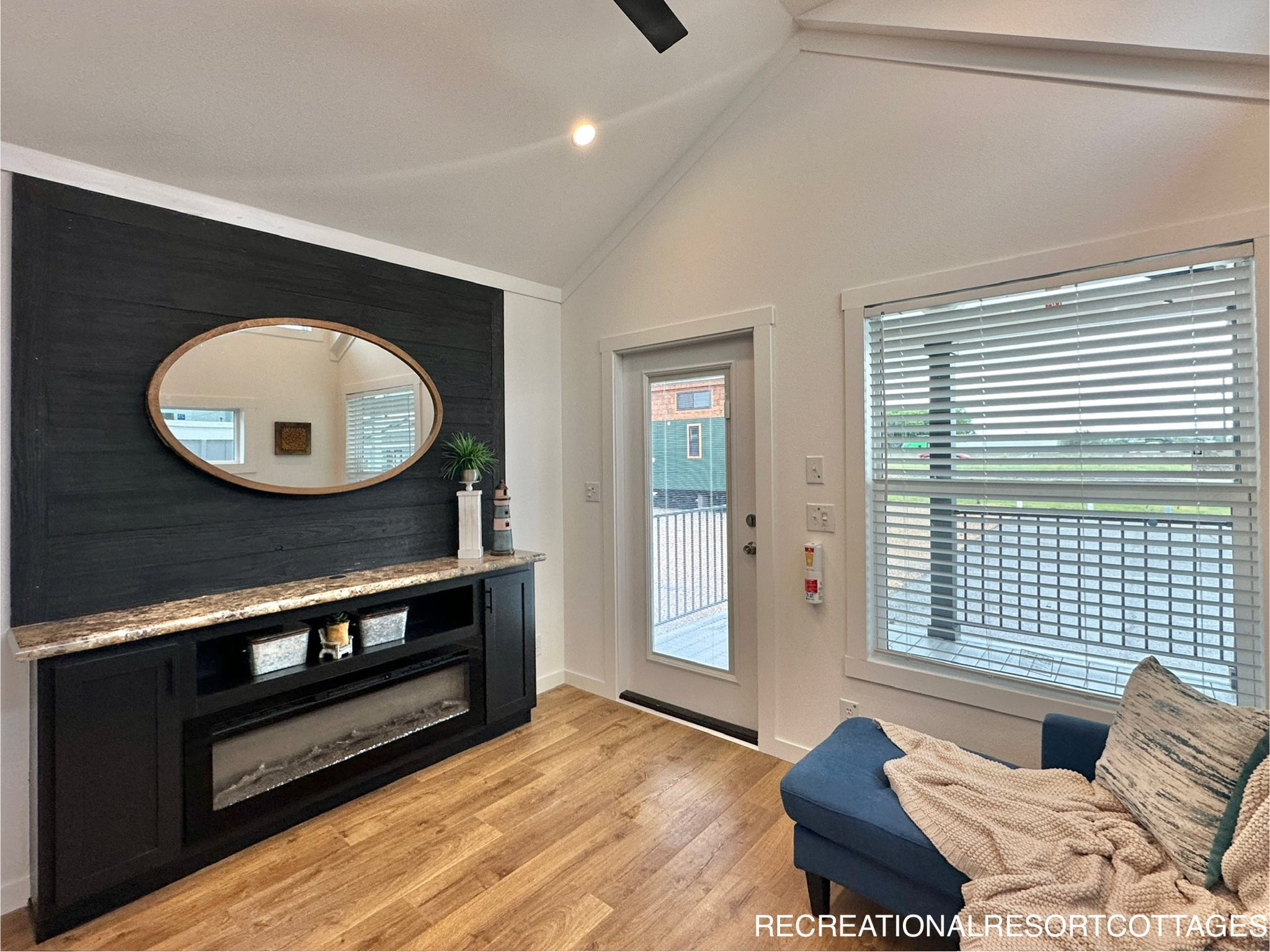 Cozy living room with a dark wood accent wall featuring a round mirror above an electric fireplace. Glass door and window with blinds on the right.