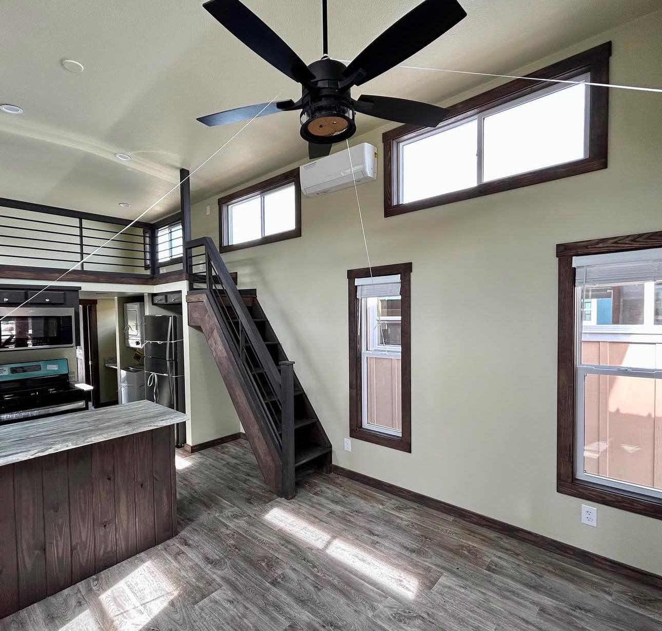 Modern tiny home interior with loft, featuring a wooden staircase, ceiling fan, and large windows. The kitchen has dark wood cabinets and stainless appliances.