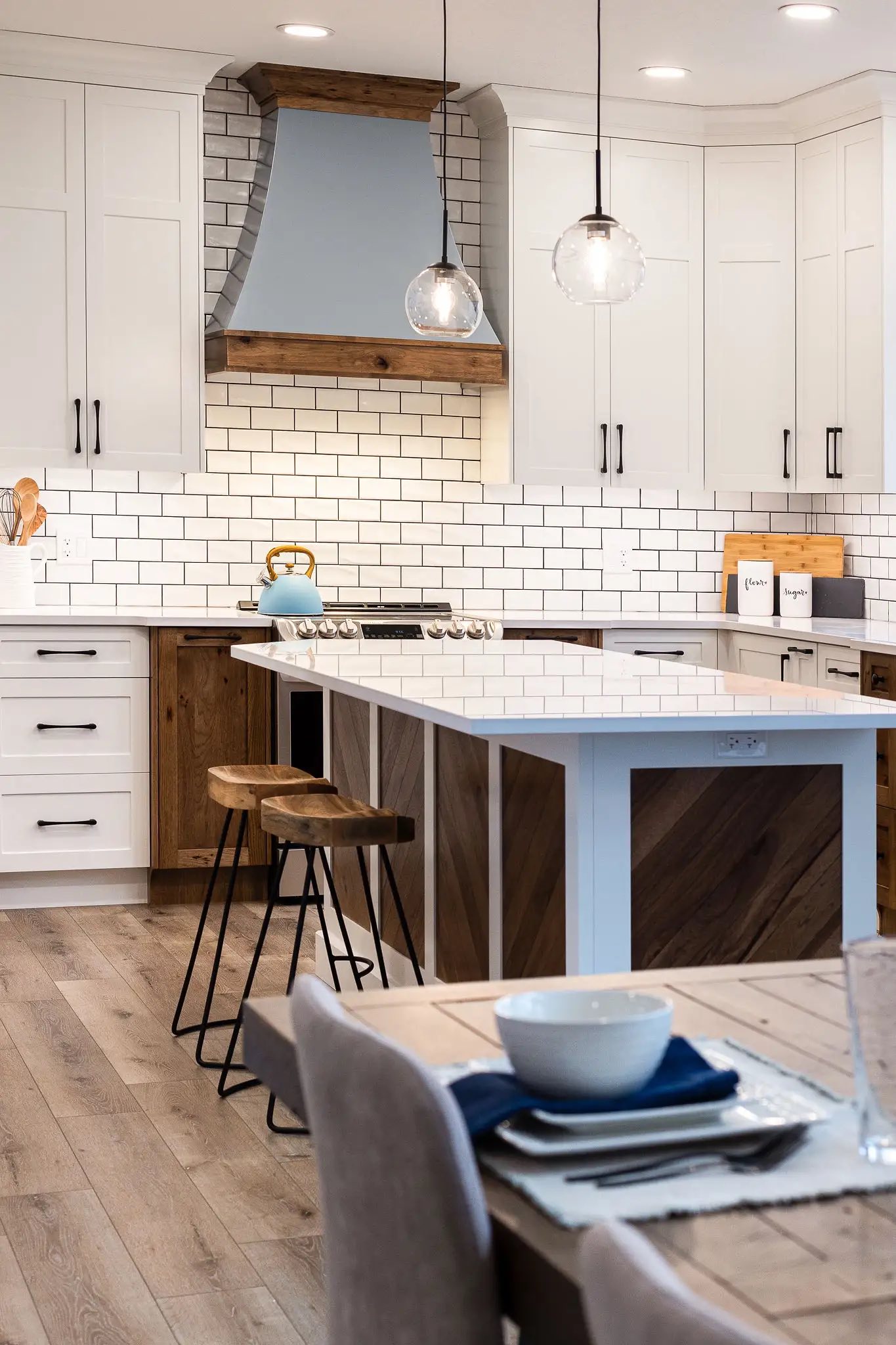 A modern kitchen with white cabinets, a blue kettle on the stove, and wooden stools at a central island. Bright and inviting atmosphere.