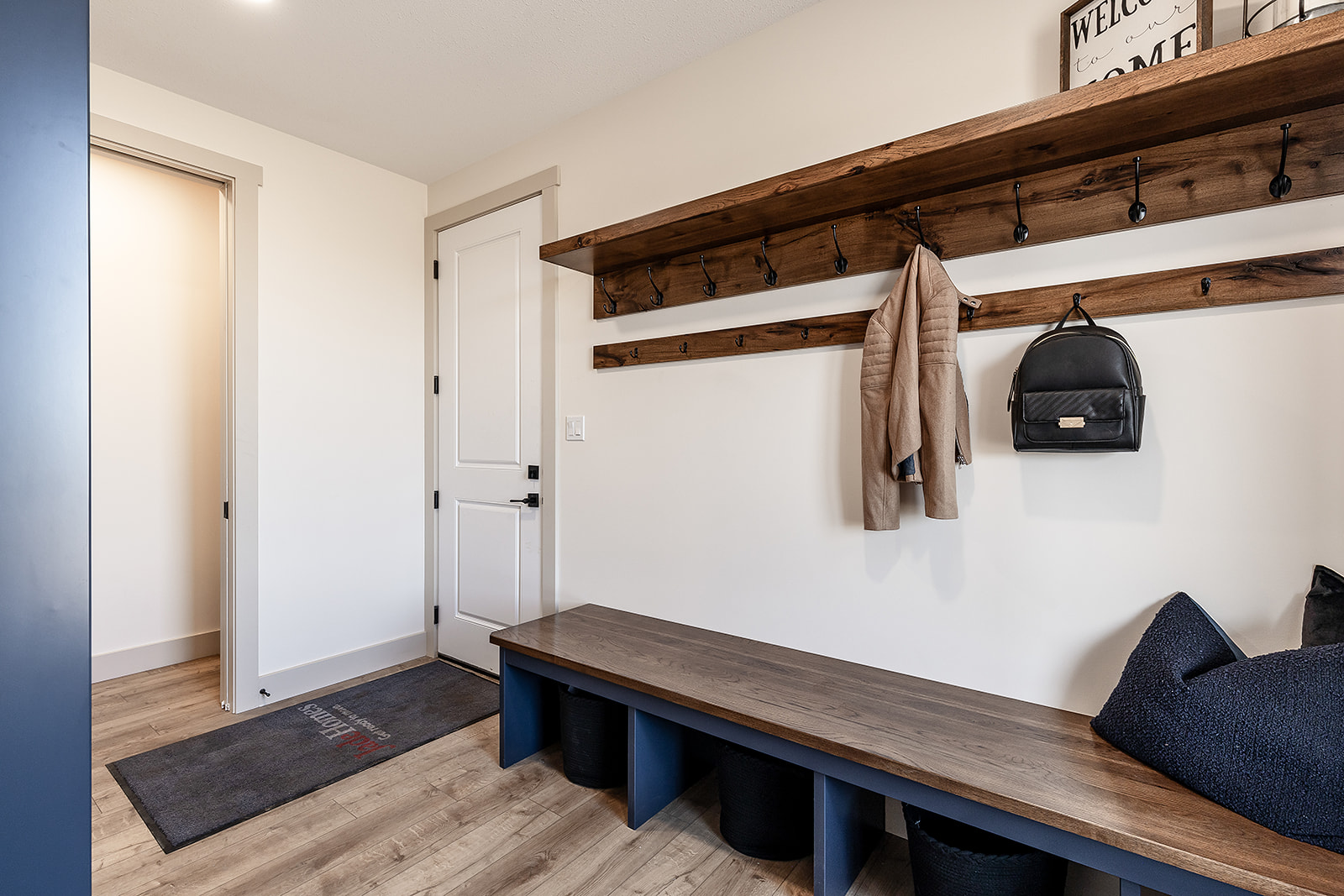A tidy mudroom features a wooden bench with cubbies, hooks on the wall holding a coat and backpack, light wood flooring, and a "Welcome" sign above.