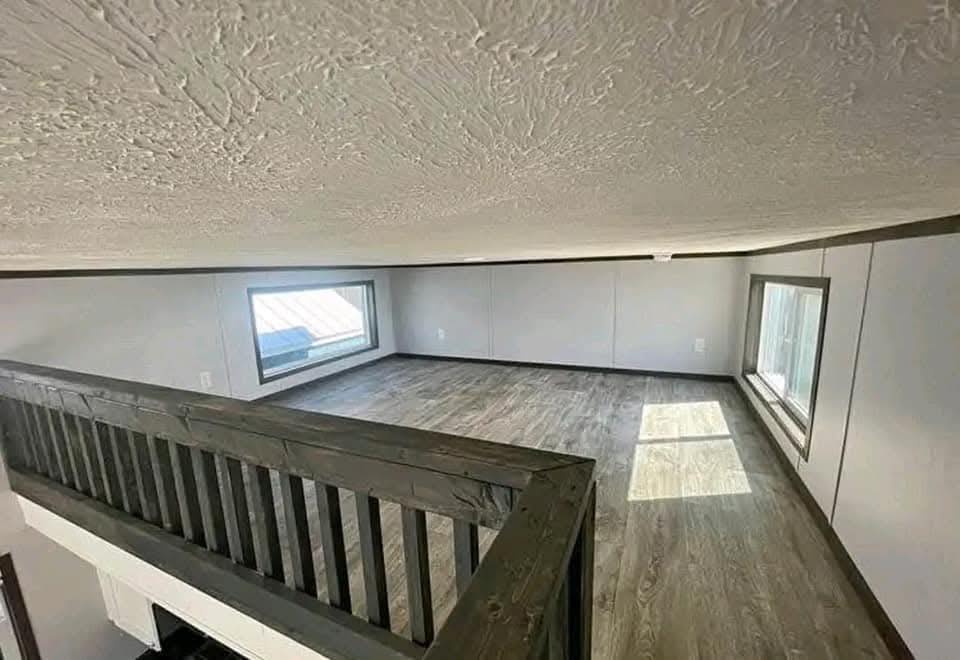 A small, minimalistic loft space with low ceilings, wooden flooring, and natural light from two narrow windows. The tone is cozy and compact.