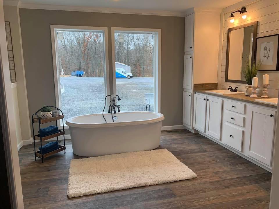 Spacious bathroom with a freestanding tub on a white rug, hardwood floor, and large windows showing trees outside. Bright, cozy ambiance.