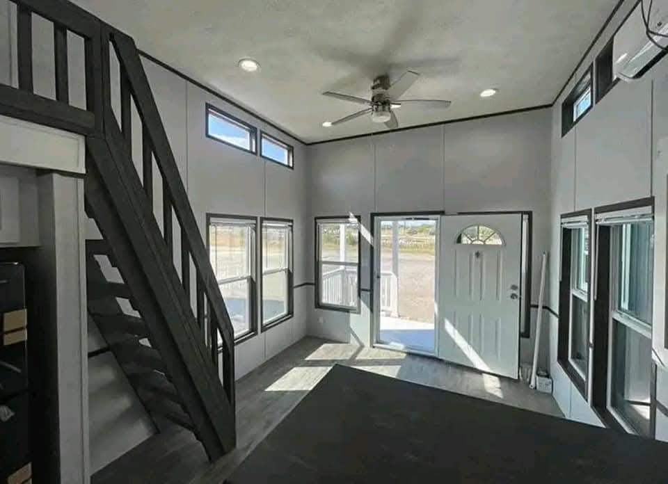 Modern tiny home interior with high ceilings, large windows, and a ceiling fan. Dark wood staircase leads to a loft, and the open door shows a bright outside.