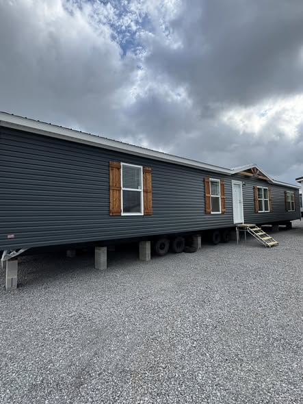 A dark gray mobile home with wooden shutters and a small staircase stands on blocks under a cloudy sky, conveying a temporary, transitional feel.