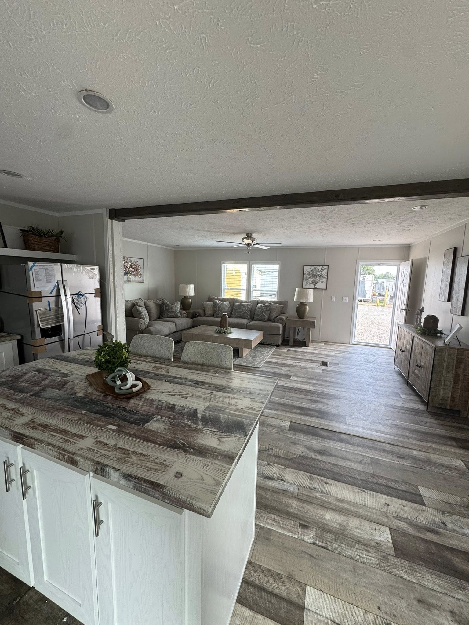 Open living area with rustic decor, featuring a wooden floor, a large sectional sofa, coffee table, and two windows. A kitchen counter is in the foreground.