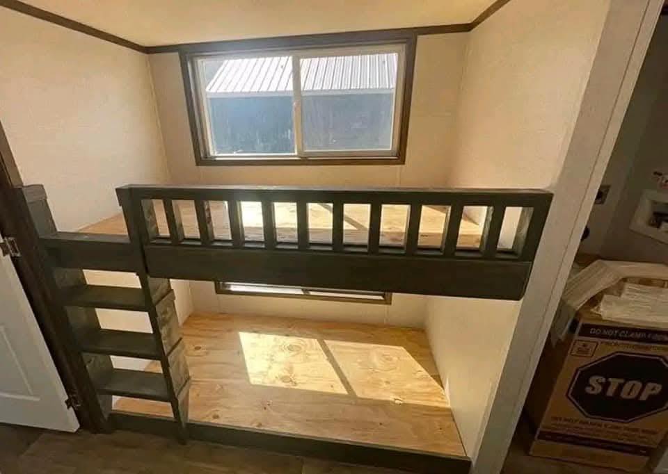 A small, well-lit room features a compact bunk bed structure with a dark wooden frame. Sunlight filters through a window, creating a warm atmosphere.