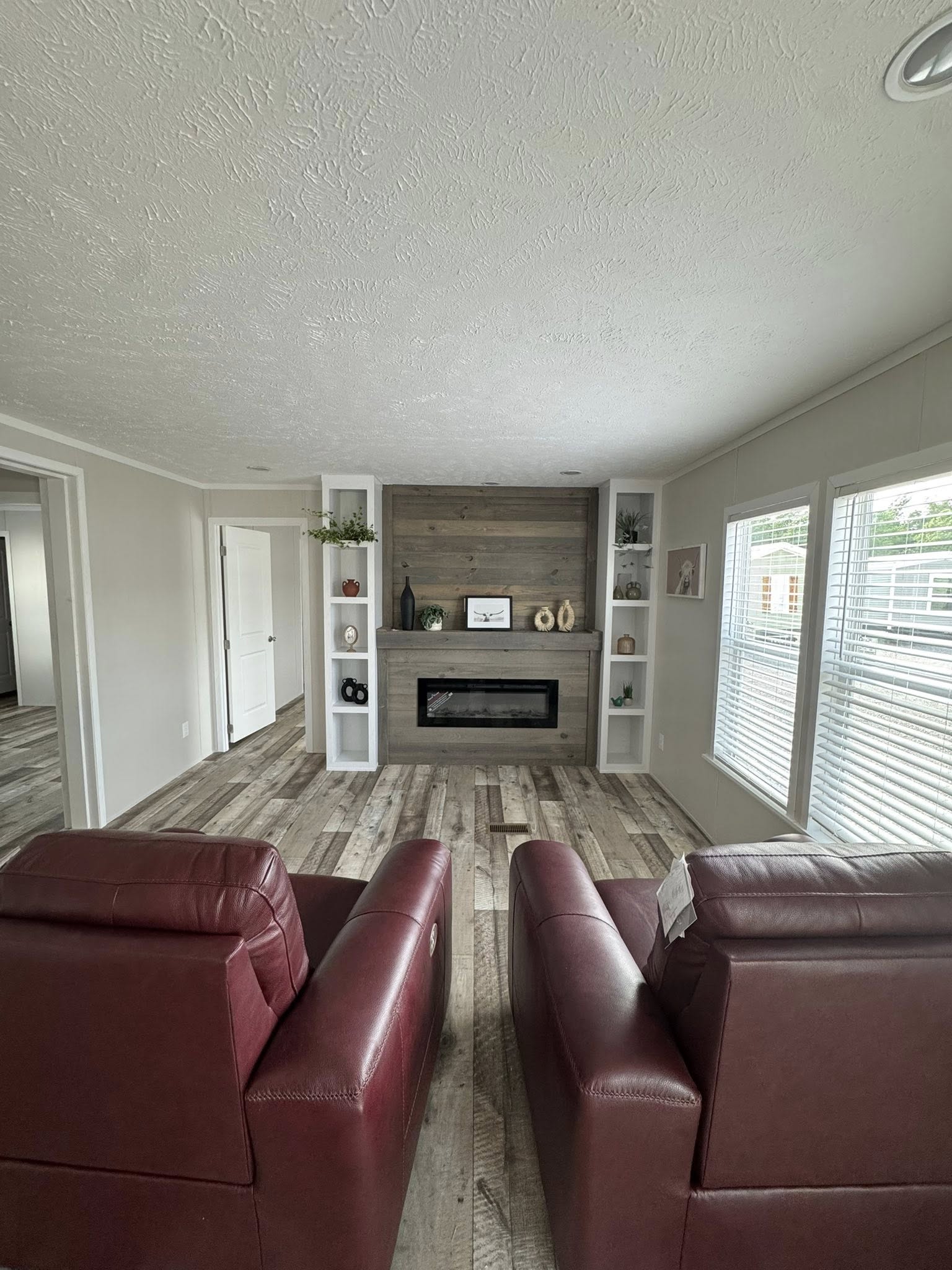 A cozy living room with two red chairs facing a modern fireplace framed by wood paneling. Shelves with decor items flank the fireplace, and large windows line the right wall, letting in natural light.