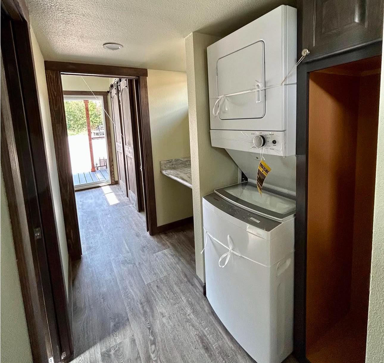 Narrow hallway with wood flooring, featuring a stacked washer and dryer on the right. At the end, a glass door opens to a sunlit porch.