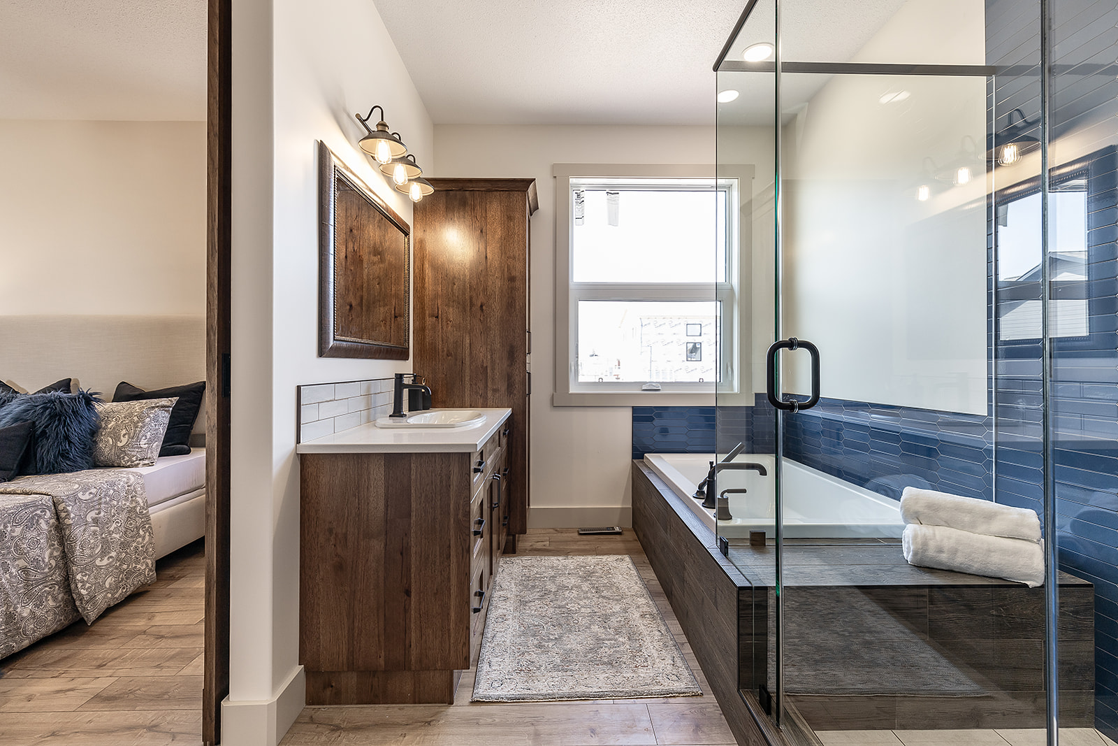 Modern bathroom with glass-enclosed bathtub and dark wood cabinets. Soft lighting and a window create a warm, inviting atmosphere. Bedroom glimpsed to the left.