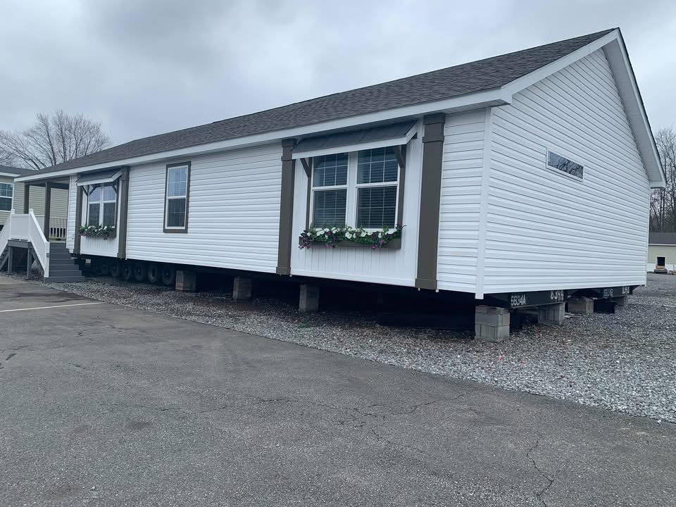A white manufactured home with a gabled roof sits on concrete blocks. The front features large windows with flower boxes. Overcast skies overhead.