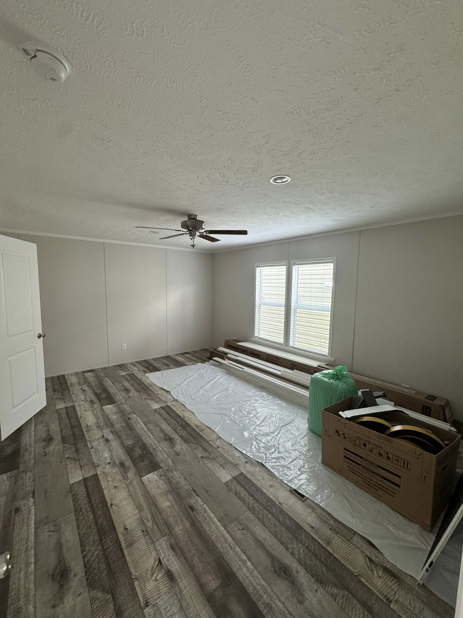 Empty room with wood-patterned flooring, beige walls, and a ceiling fan. Two windows allow natural light. Flooring materials and boxes are on the floor.