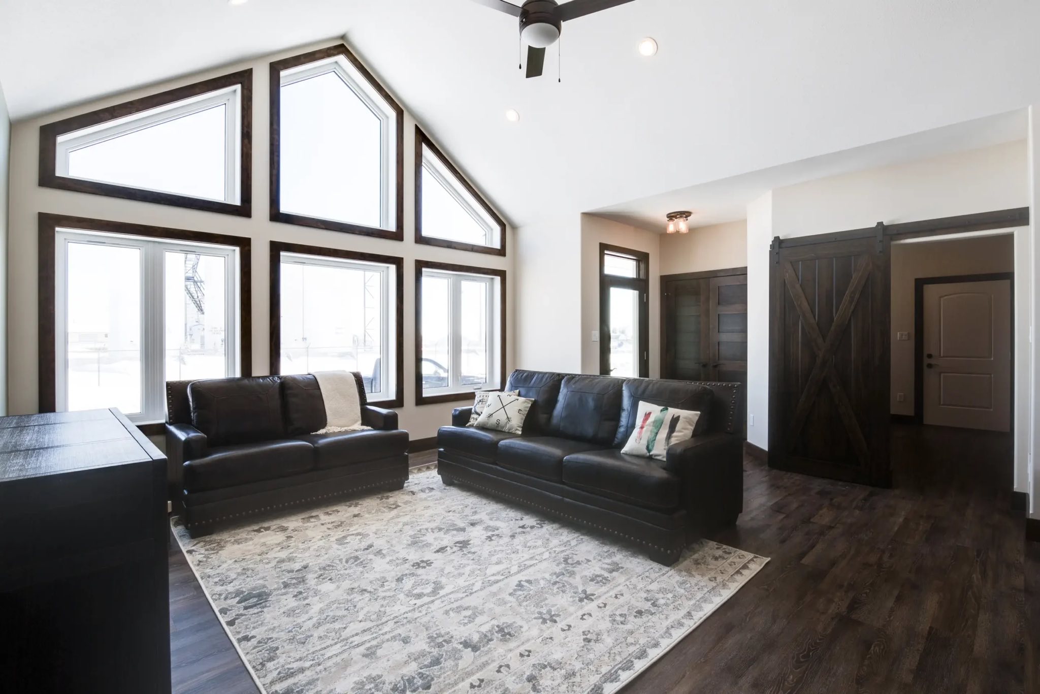 Bright living room with high vaulted ceiling and large triangular windows. Features dark leather sofas on a patterned rug, and a rustic barn door.
