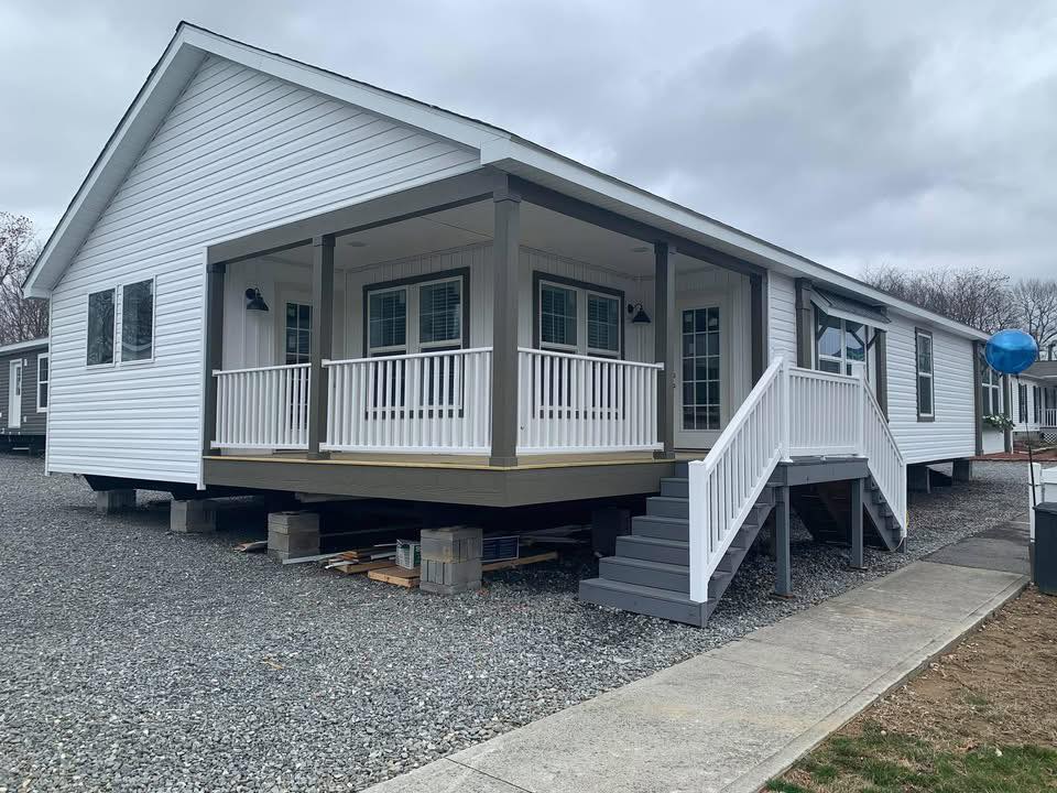 A white modular home with a spacious porch, white railings, and gray steps. It rests on concrete blocks over a gravel lot, under an overcast sky.
