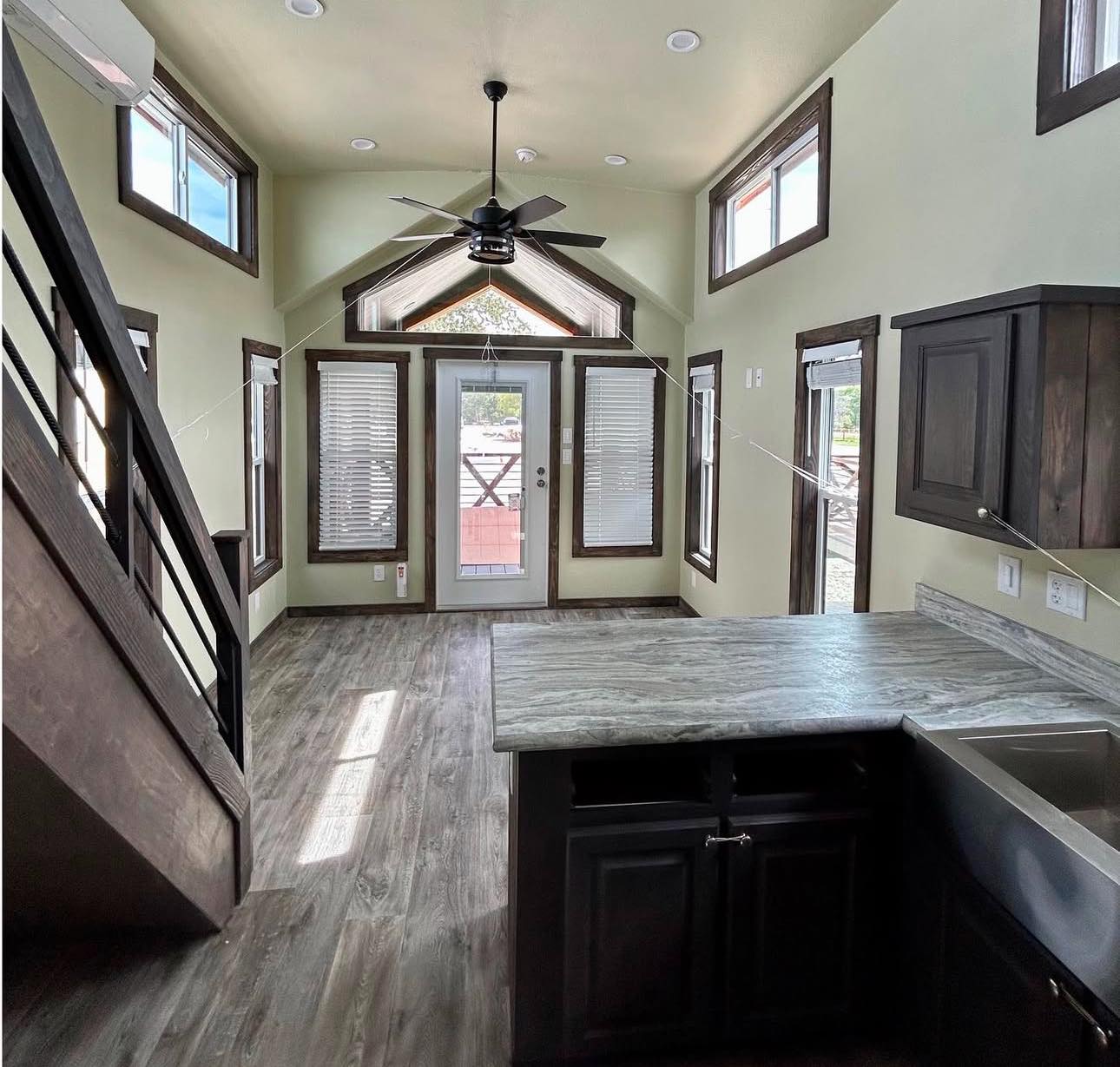 Modern tiny house interior with high ceilings, large windows, and wooden floors. A fan hangs above and light floods in, creating an airy feel.