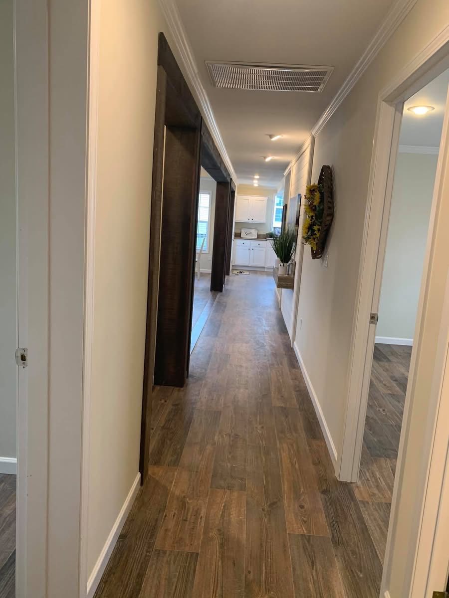 A bright hallway with wooden flooring leads to a kitchen with white cabinets. Sunflowers hang on a wall, and a plant is in the corner, creating a welcoming vibe.
