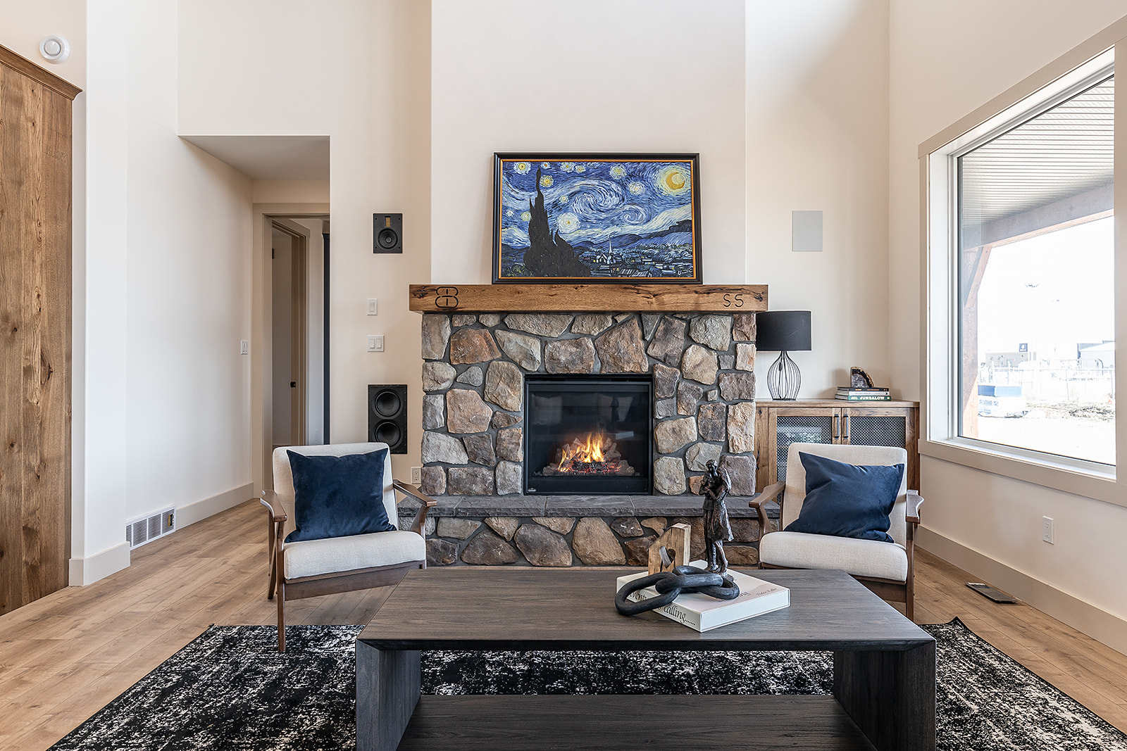 Cozy living room with a stone fireplace, two armchairs with navy cushions, and a modern coffee table. A "Starry Night" painting hangs above.