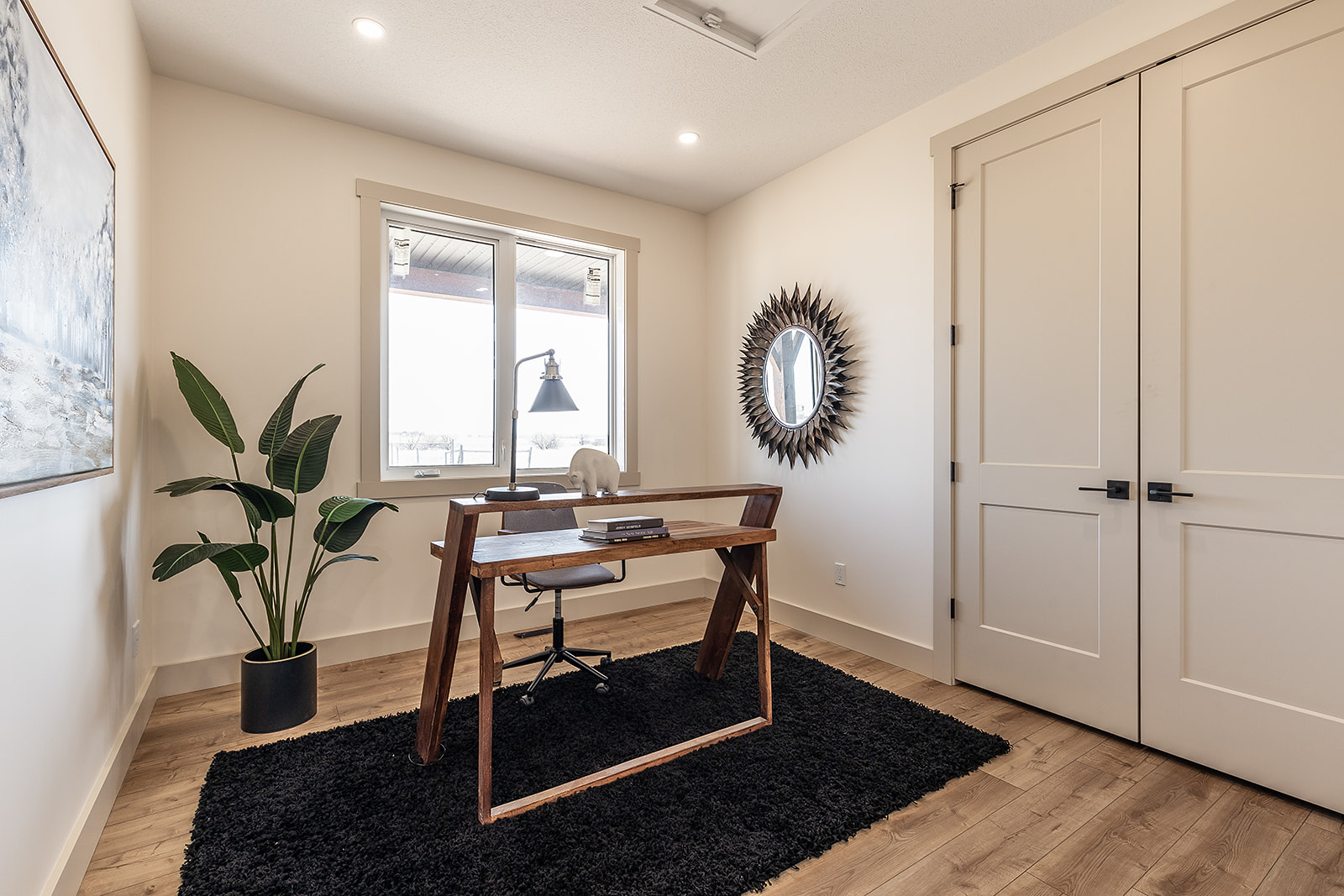 Minimalist home office with a wooden desk on a black rug, swivel chair, and a lamp. A large plant and sunburst mirror enhance the decor. Calm atmosphere.