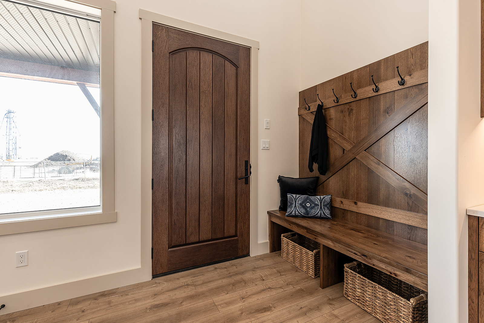 Cozy entryway with wooden door and bench. Coat hooks and woven baskets add functionality. Natural light through window. Warm and inviting tone.