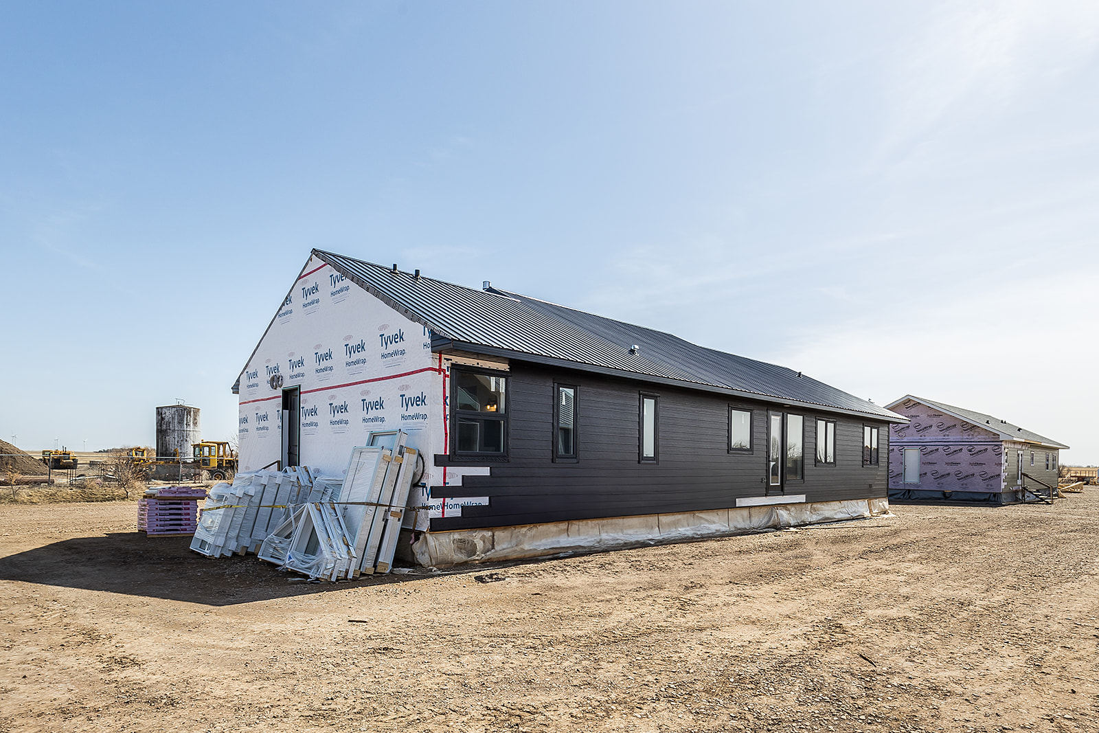A partially constructed modern house sits on a dirt lot under a clear blue sky. The building has dark siding, unfinished ends, and materials stacked nearby.