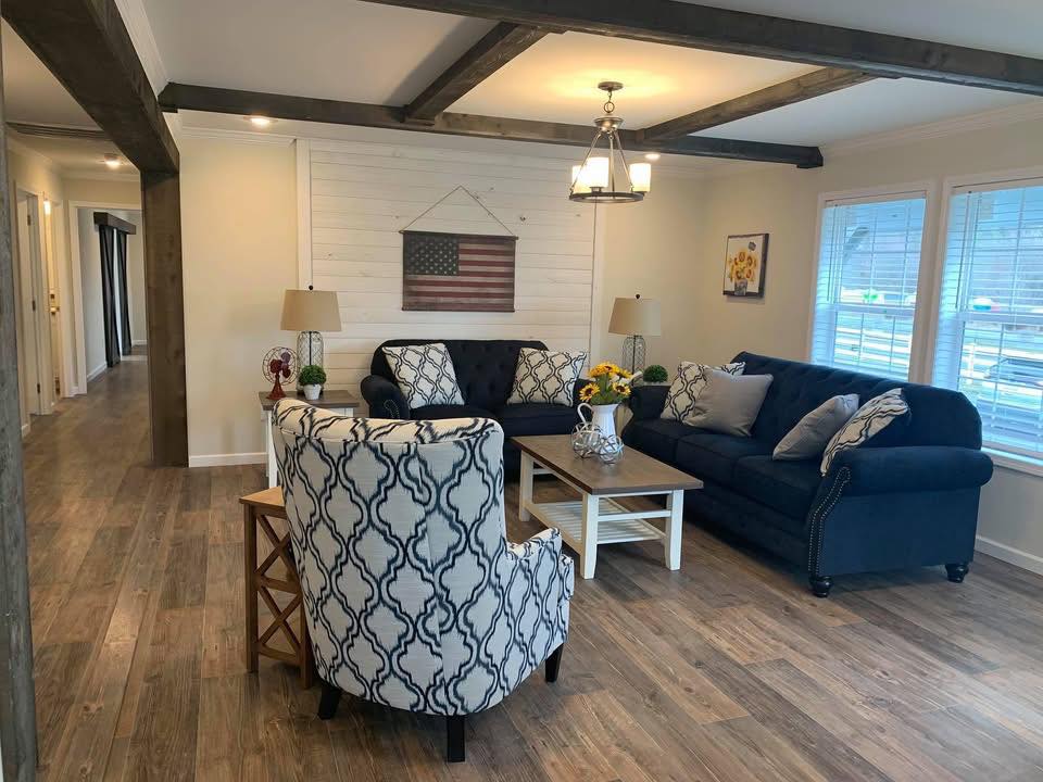 Cozy living room with navy sofas, patterned armchair, and wooden coffee table. American flag art on the wall; bright with natural light from windows.