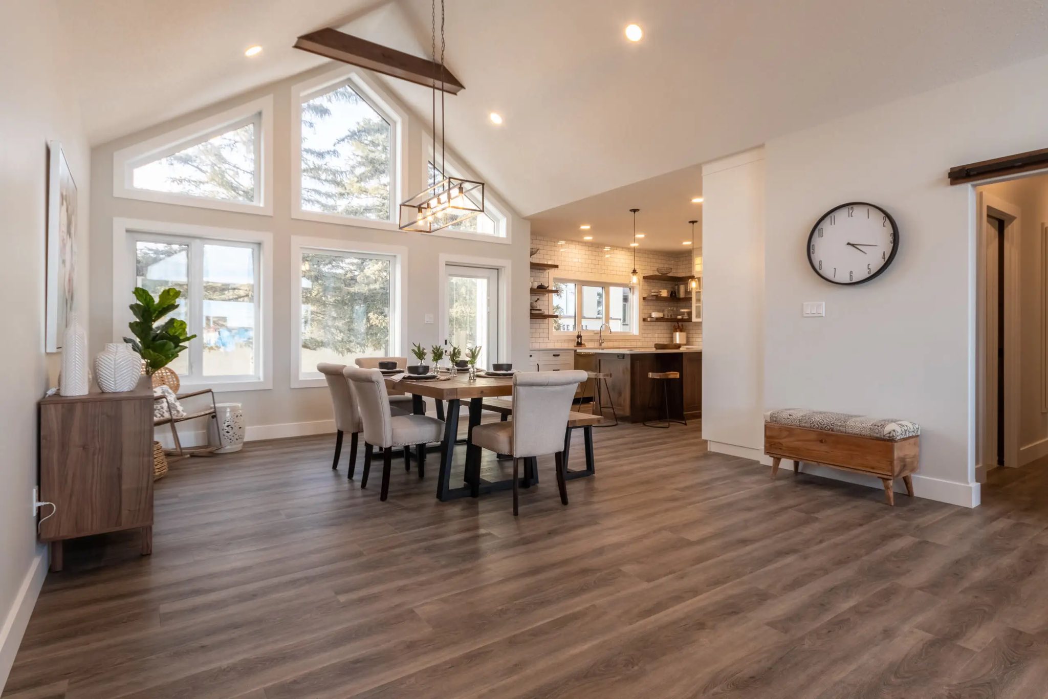 Spacious, modern dining room with vaulted ceiling and large windows. Wooden table with six chairs, pendant light, and open kitchen in the background.