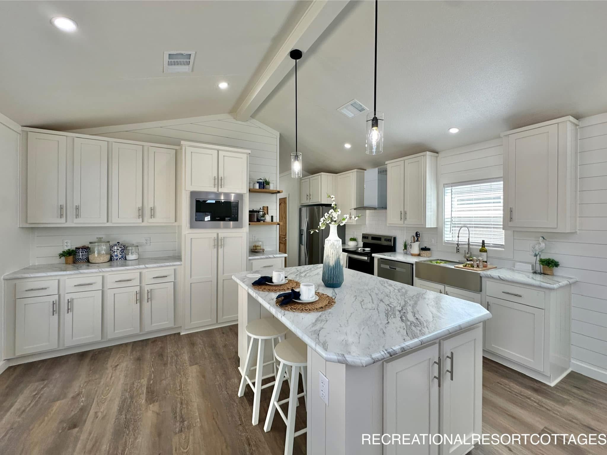 Spacious kitchen with white cabinets, marble countertops, and a central island. Wooden floor, pendant lights, and a vase add a modern touch.