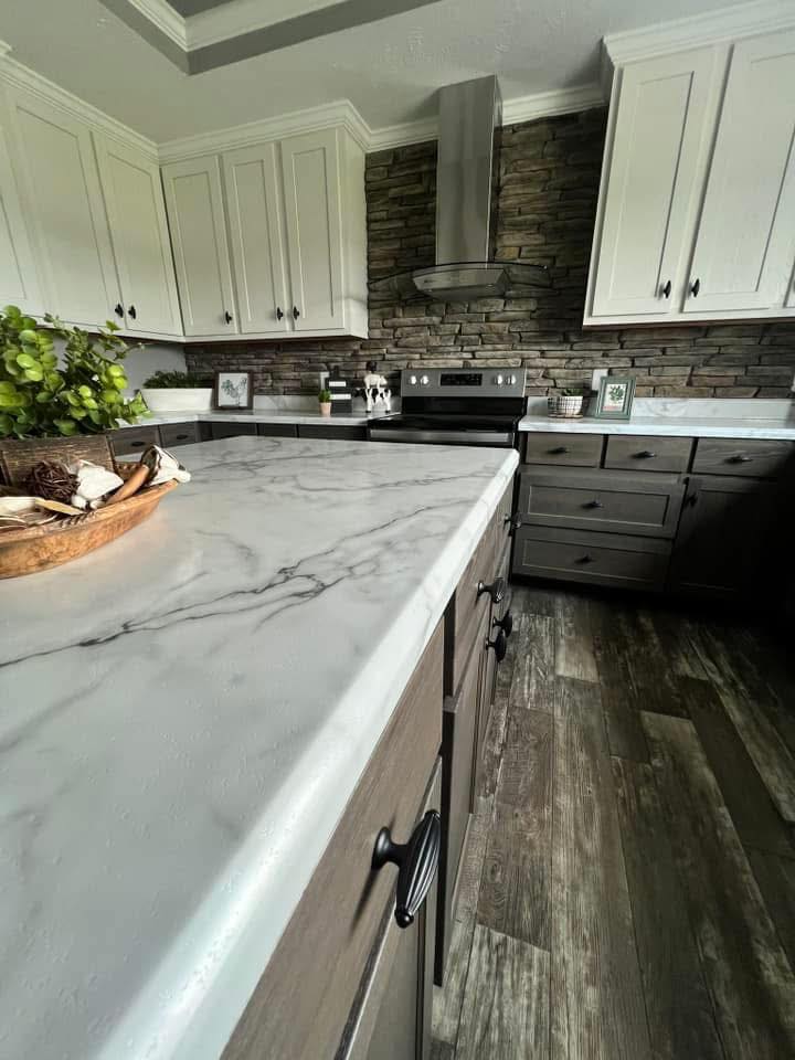 Modern kitchen with a marble countertop, dark wood cabinets, white upper cabinets, and a stainless steel stove against a textured stone wall. Cozy yet sleek ambiance.