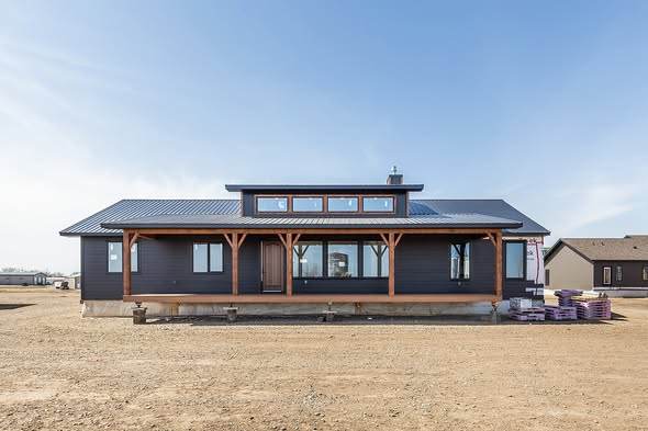 Single-story black house with a metal roof, large windows, and wooden accents on an open dirt lot. The clear sky adds a serene backdrop.