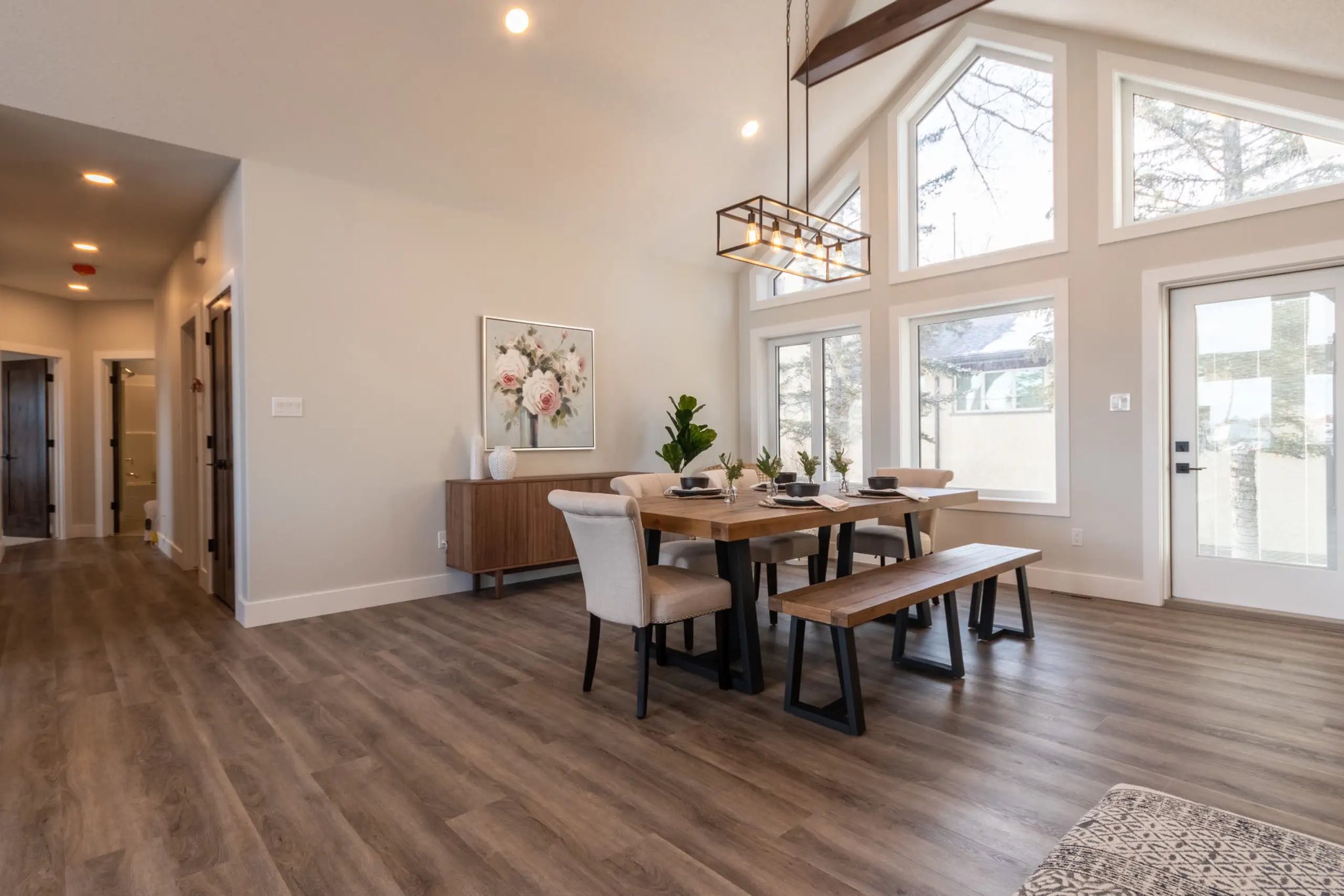 Modern dining room with vaulted ceiling, large windows, and natural light. Features a wooden table with chairs and bench, light fixture above. Cozy, elegant ambiance.