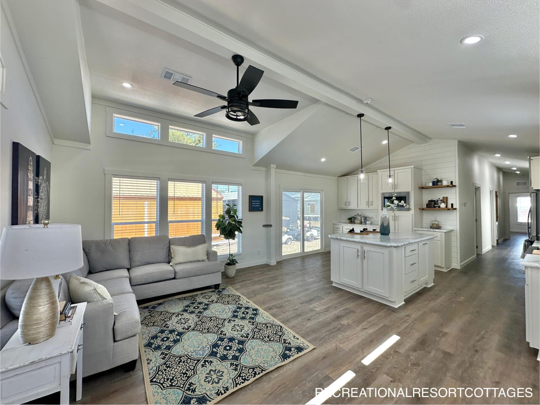 Spacious, modern living area with a high ceiling and recessed lighting. Features a ceiling fan, light gray sofa, decorative rug, and an open kitchen with white cabinets and island. Natural light streams through large windows, creating an inviting atmosphere.