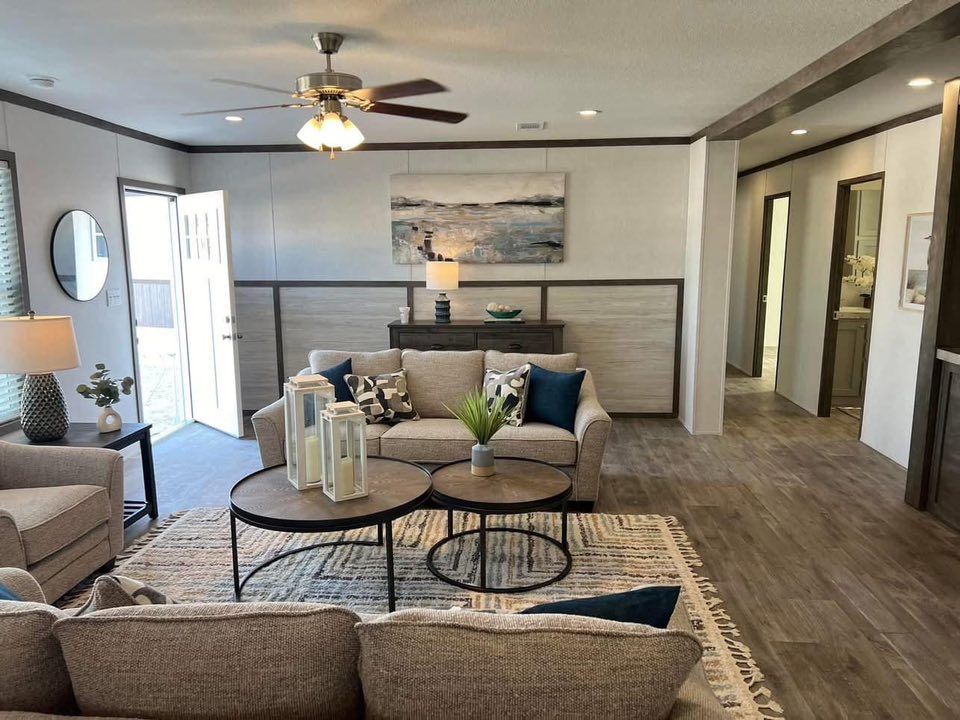Cozy living room with neutral tones, featuring beige sofas, circular coffee tables, and a textured rug. Open door lets in light, creating a warm atmosphere.