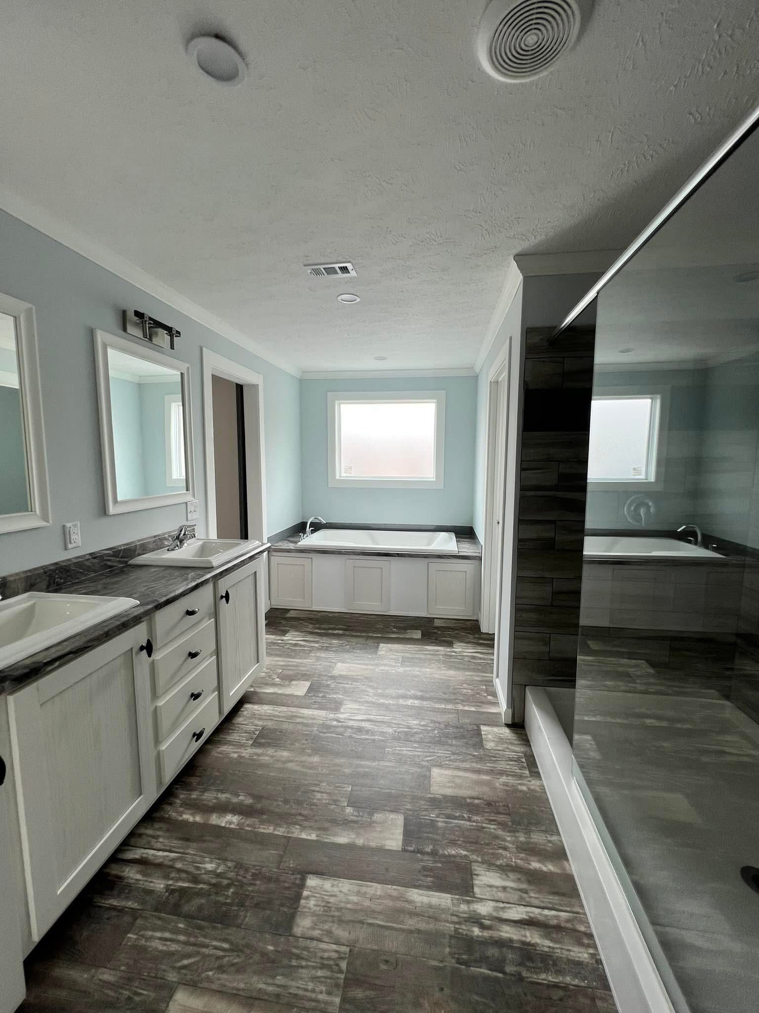 Modern bathroom with light blue walls, featuring a large bathtub under a window, dual sinks with marble countertops, and a glass-enclosed shower.