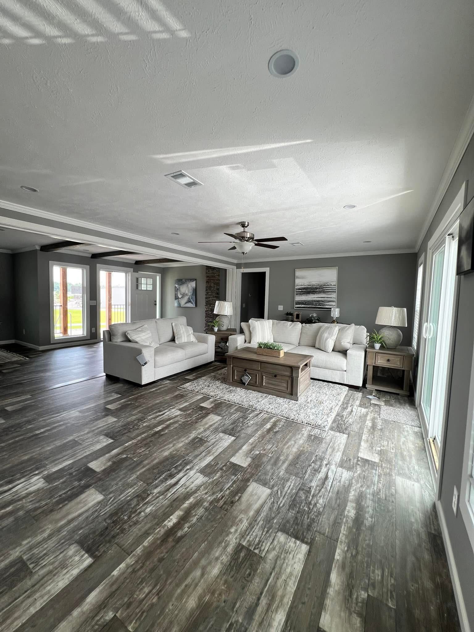 Spacious, modern living room with gray walls, wood floor, white sofas, wooden coffee table, and a ceiling fan. Sunlight filters through large windows.