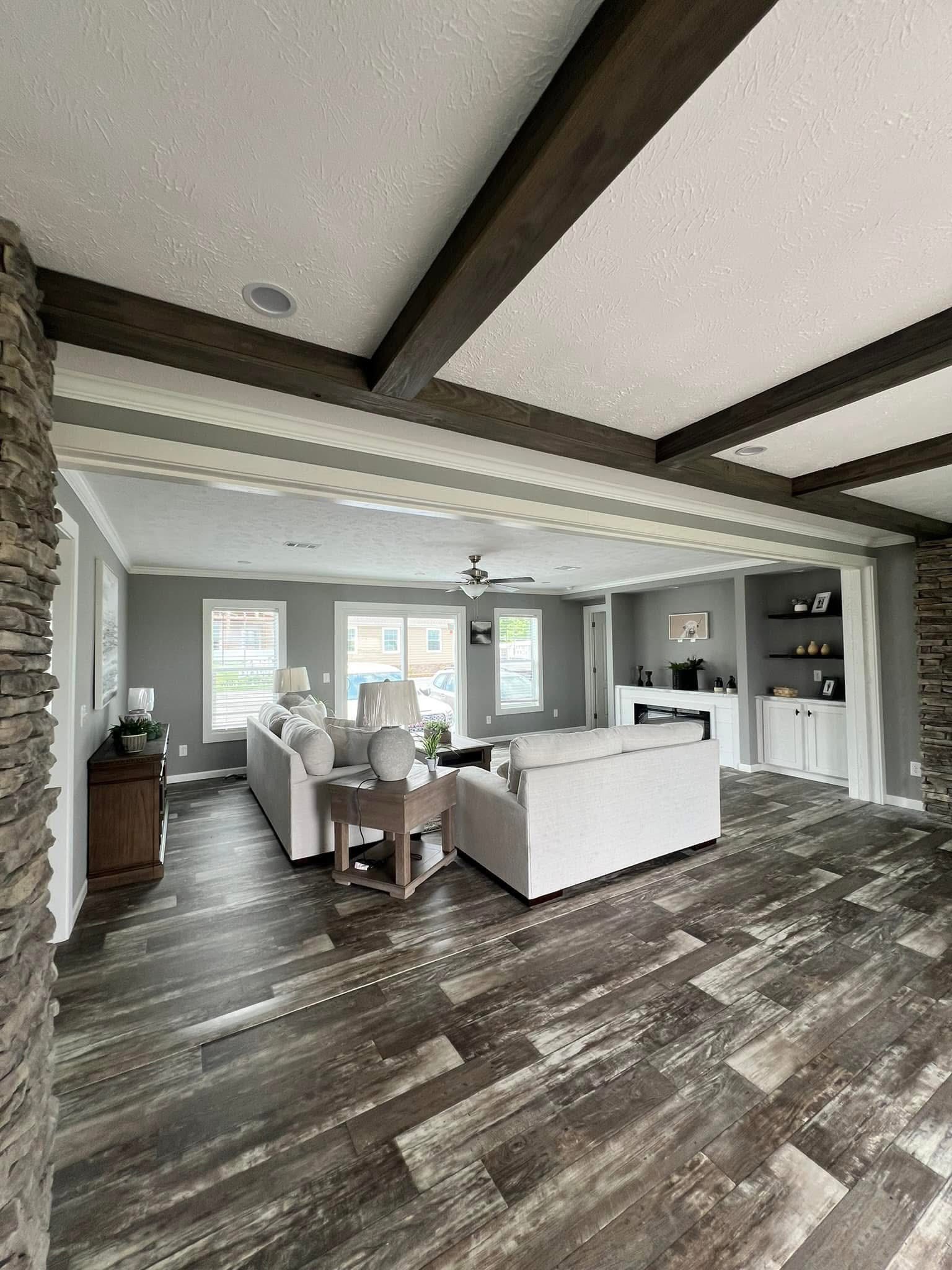 Spacious modern living room with gray walls, wood ceiling beams, and brown hardwood floors. Features white sofas, a coffee table, and built-in shelves.