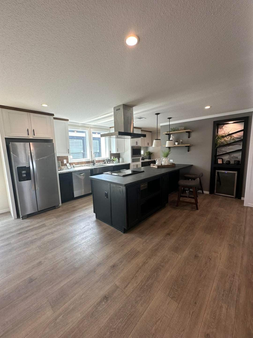 Modern kitchen with a large black island, light wood floors, stainless steel appliances, and white cabinets. Open shelves hold plants, creating a cozy feel.