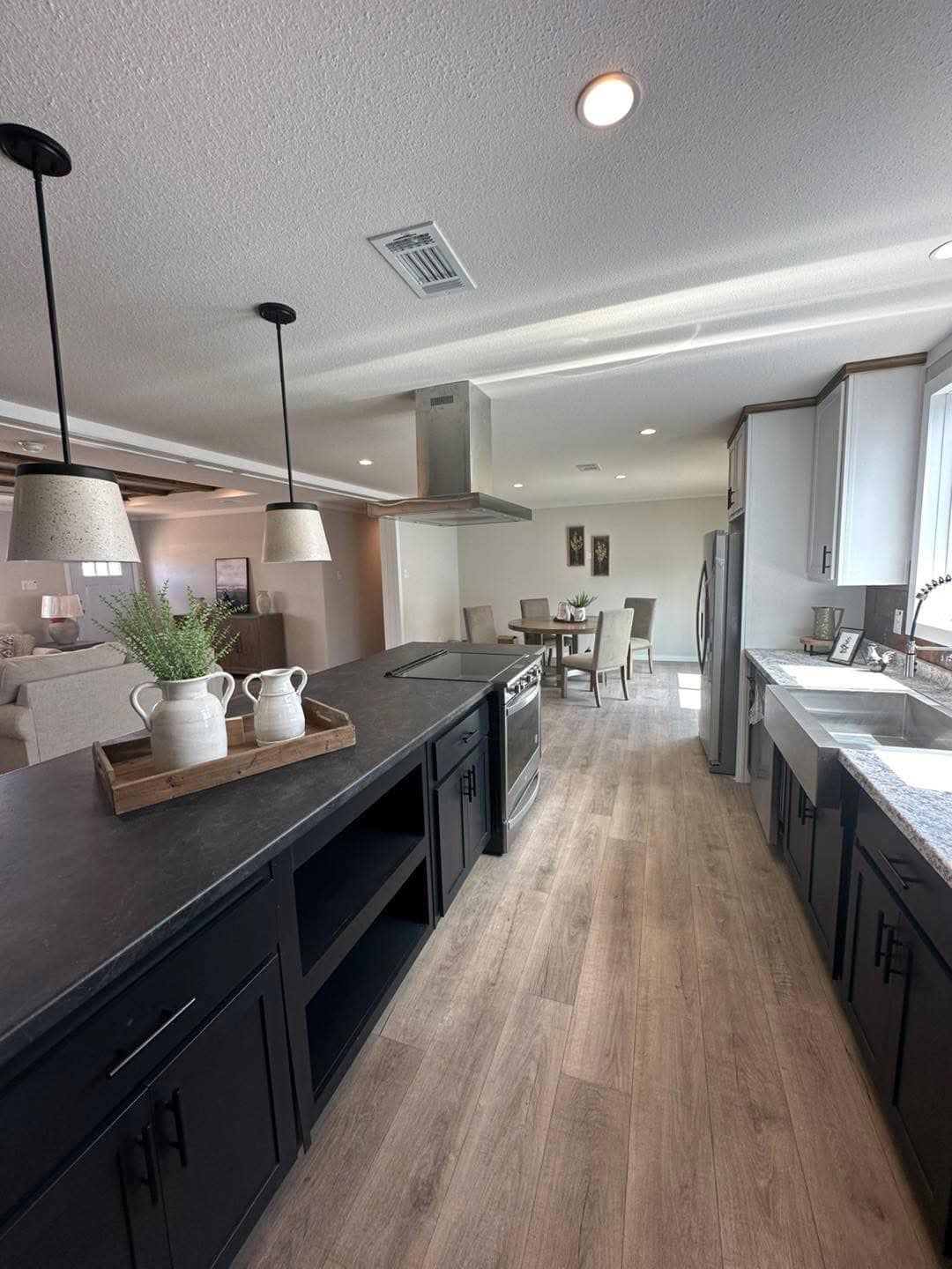 Modern kitchen with dark cabinetry, light wood flooring, and stainless steel appliances. A farmhouse-style island holds decorative white pitchers.