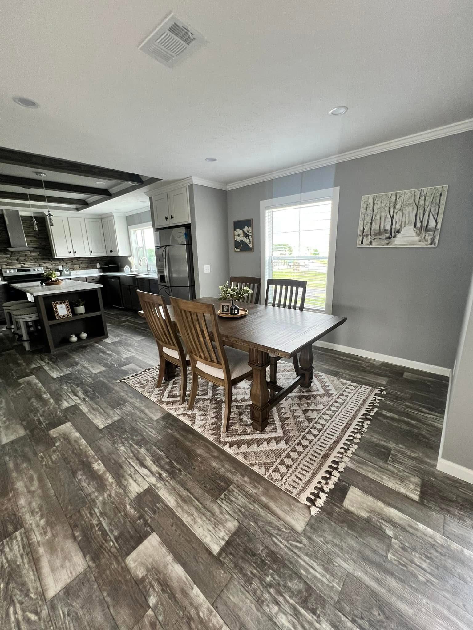 Spacious kitchen and dining area with gray walls and wood flooring. A wooden table with four chairs sits on a patterned rug, near large windows. Modern and inviting ambiance.