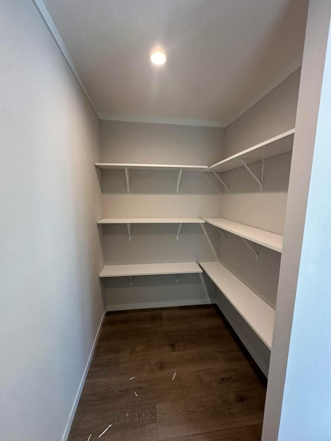 Small, empty pantry with white walls and four white shelves on gray brackets. Dark wood flooring and overhead lighting create a clean, minimalist look.