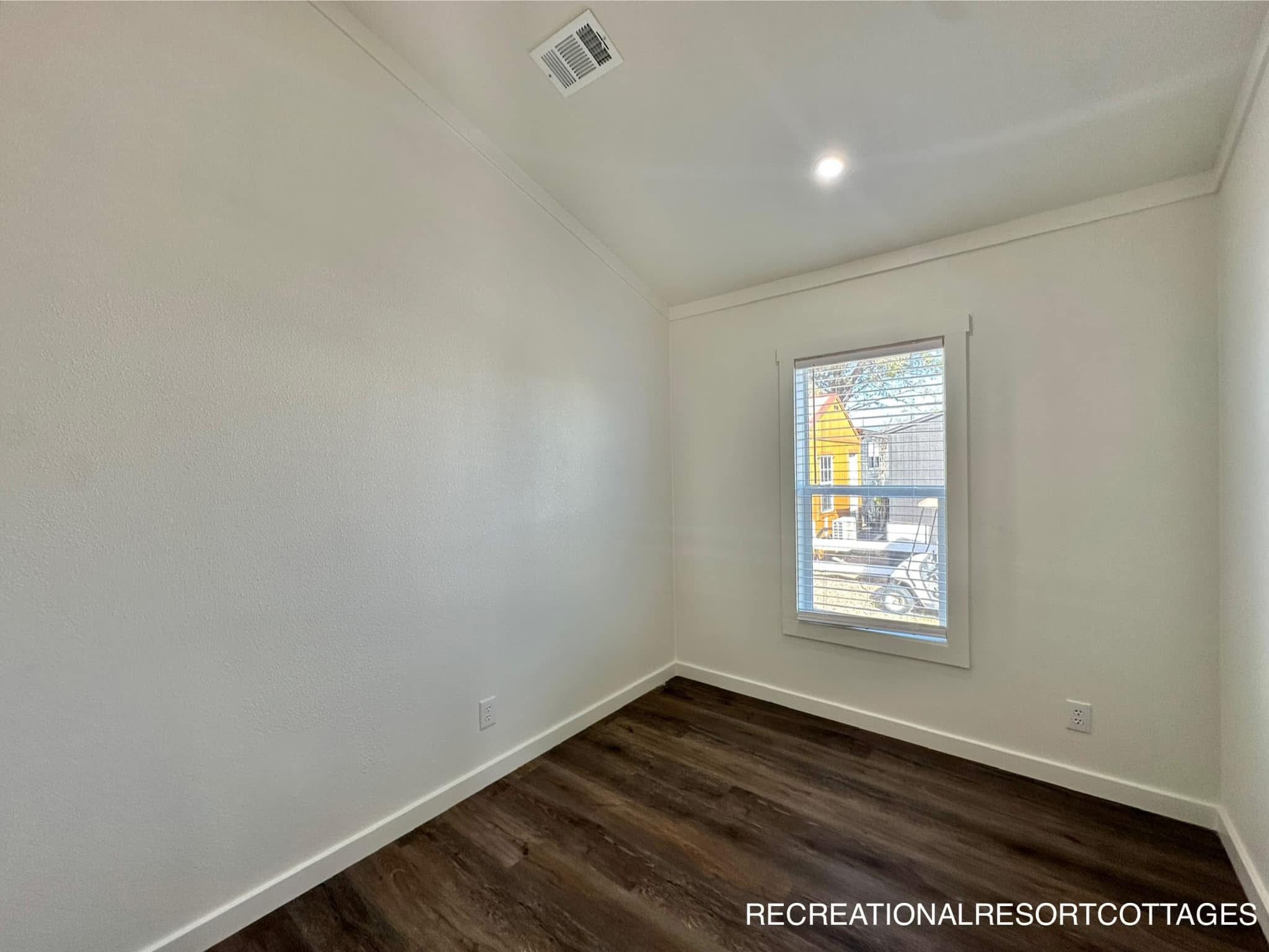 A small, unfurnished room with white walls and dark wood floors. It has a high ceiling and a window showing a glimpse of a yellow house outside.