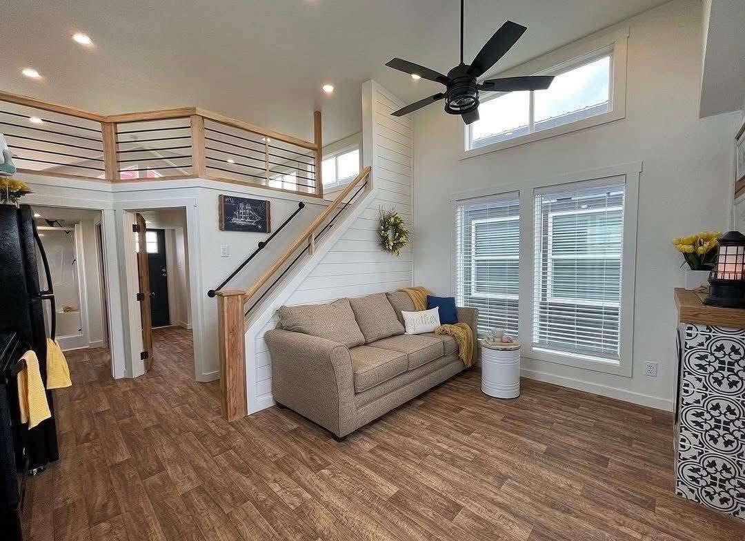 A cozy living room with wooden floors and a beige couch accented by a yellow throw and pillows. A staircase with wooden railing leads to a loft. Large windows and a ceiling fan provide a bright and airy feel.