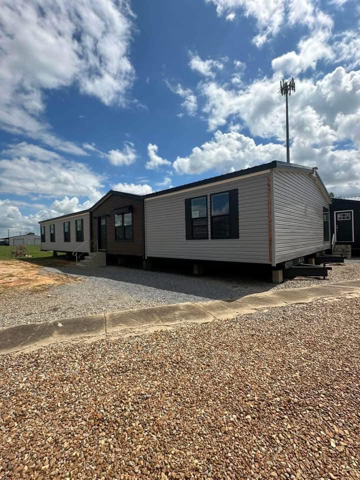 A large manufactured home sits on a gravel lot under a partly cloudy sky. The structure is light gray with dark trim, conveying a sense of simplicity and modernity.