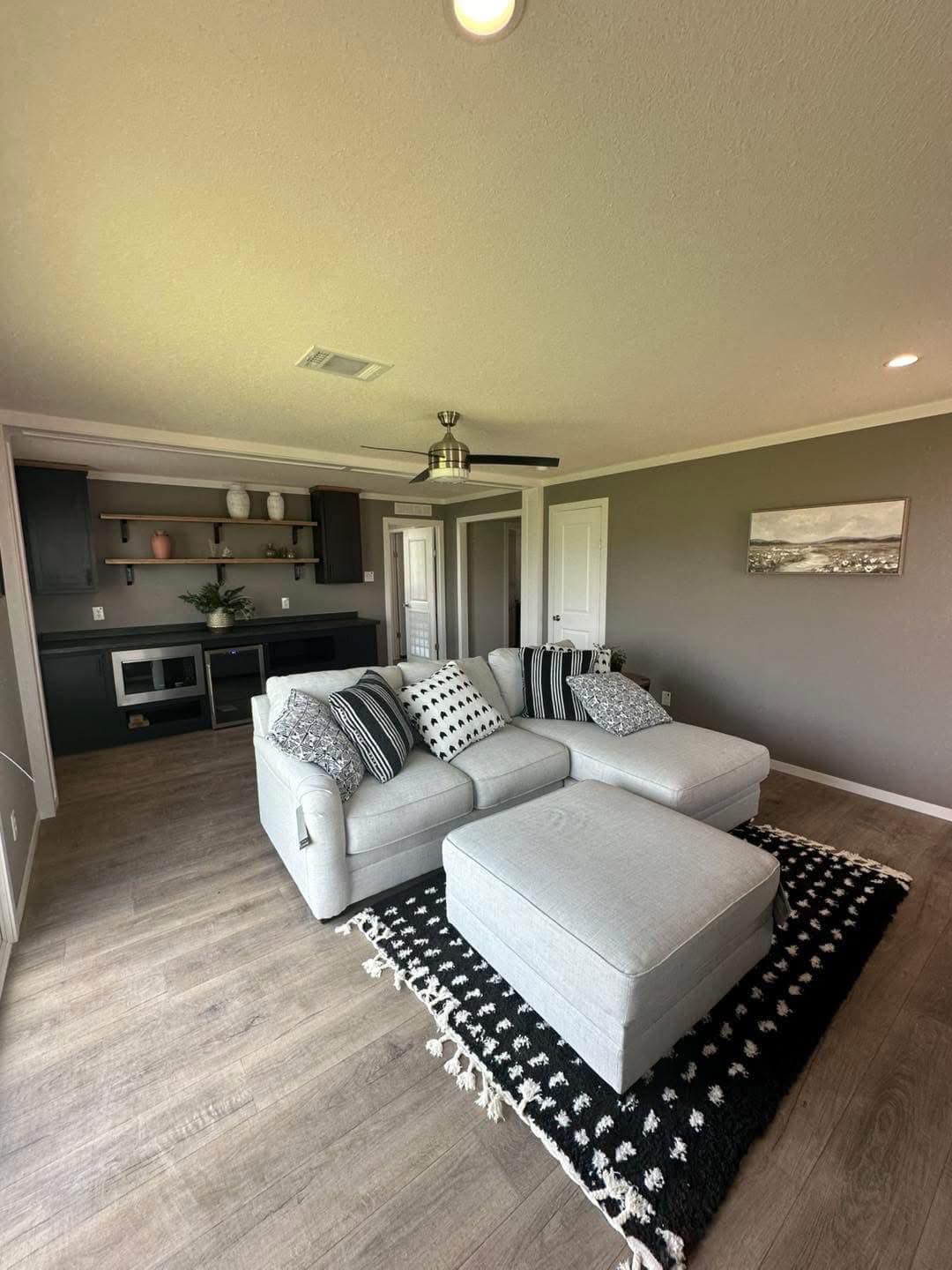 A cozy living room with a gray sectional sofa adorned with black and white patterned cushions, set on a dark rug. Modern decor includes a ceiling fan, gray walls, and a decorative shelf with plants, creating a stylish, relaxed atmosphere.