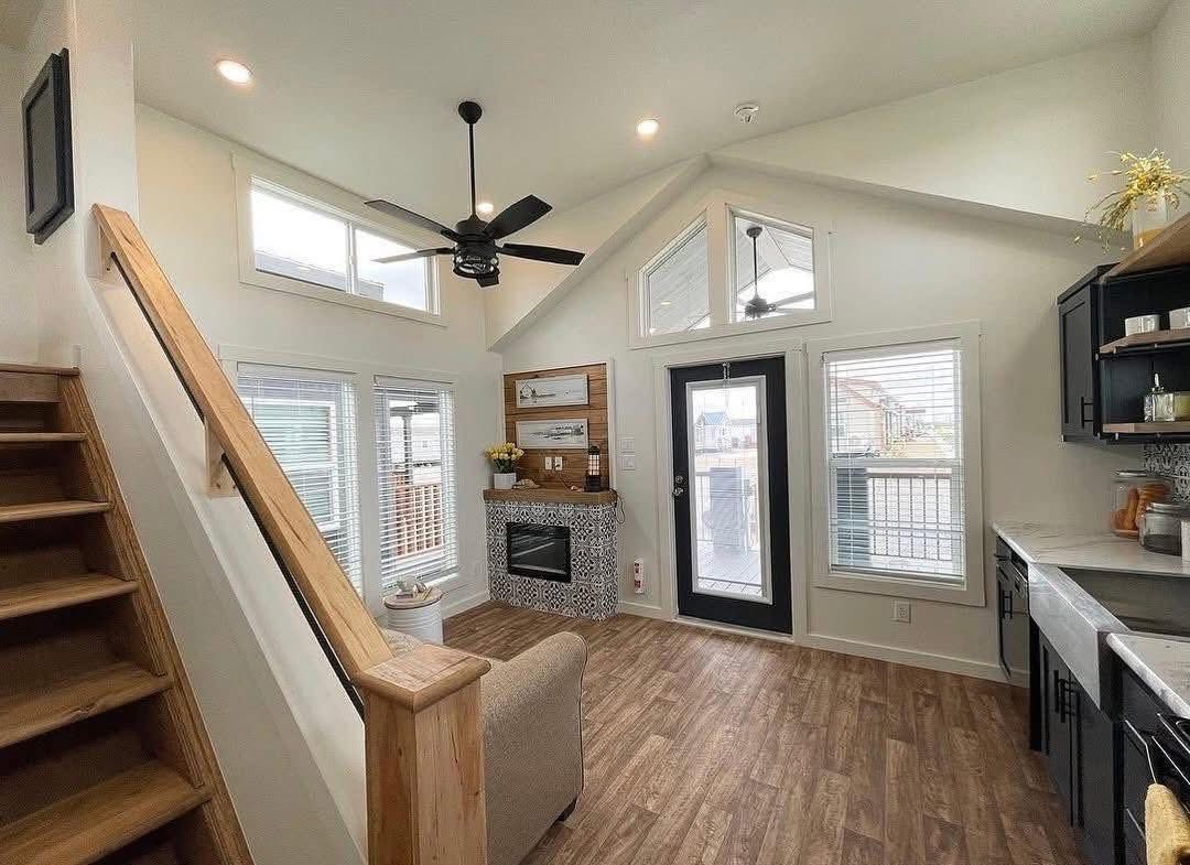 Cozy living room with high ceilings, wooden floors, and a black ceiling fan. Features a fireplace, stairs to a loft, and black-framed glass doors.