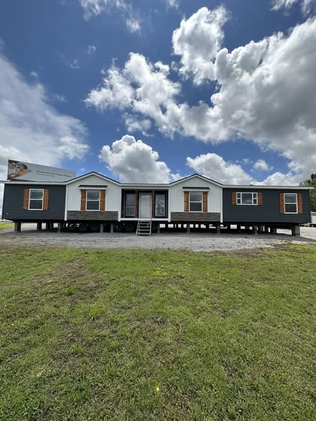A mobile home set above the ground on stilts under a partly cloudy sky. The exterior features a mix of white and dark siding, with front steps leading up.