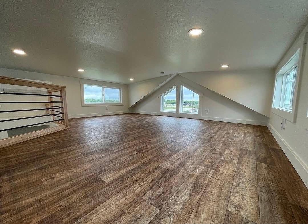 Spacious attic room with wood floor, angled ceiling, and triangular window overlooking scenic view. Bright, airy atmosphere with recessed lighting.